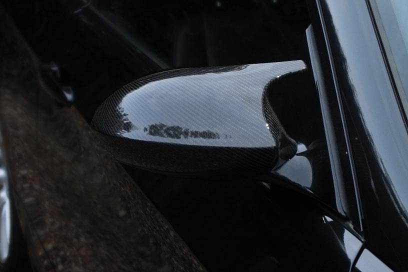 A carbon fiber side mirror reflects light on a sleek, black car parked outdoors, surrounded by a dark environment, creating a subtle contrast between the glossy mirror and matte surroundings.