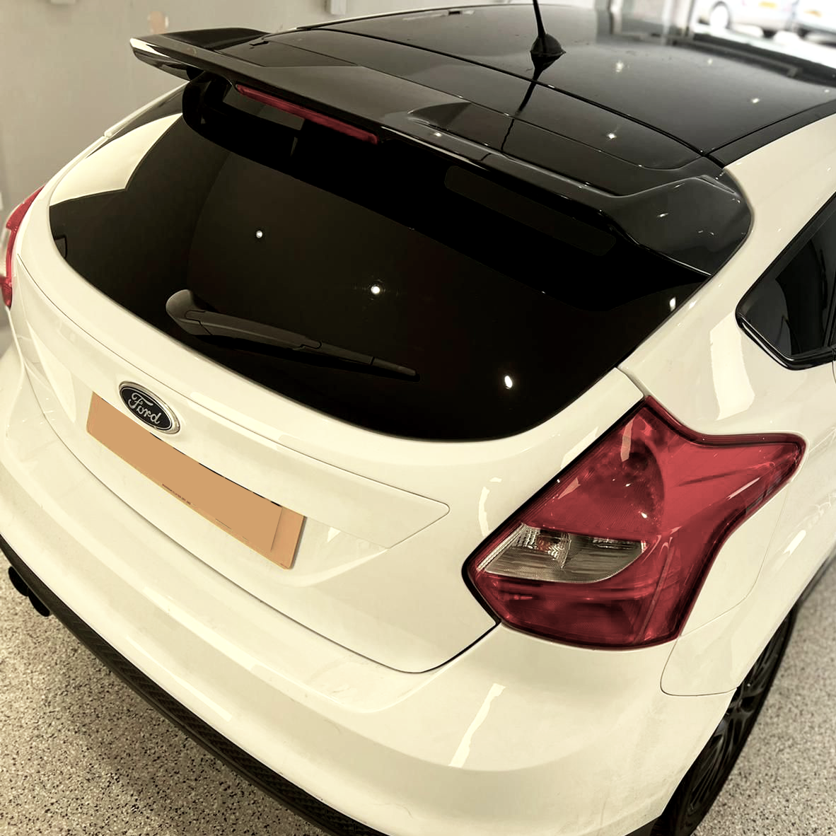 A white Ford car is parked indoors, featuring a black roof and prominent rear spoiler. It has a visible rear wiper and distinctive red tail lights against a sleek showroom environment.