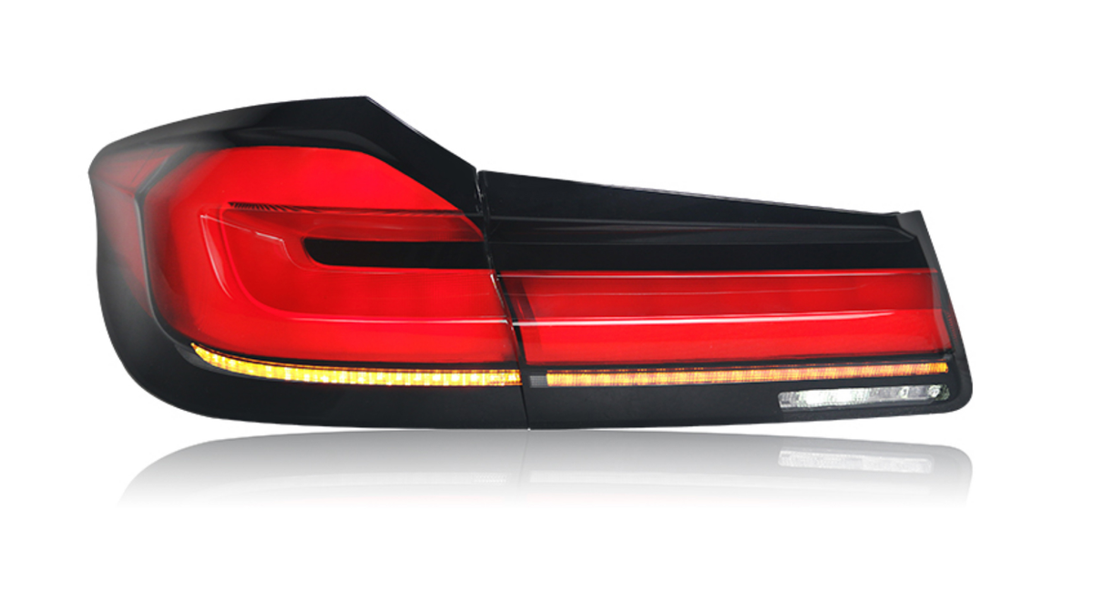 A car taillight, illuminated in red and amber, reflects on a white surface. The sleek design features multiple segments, with a glossy black frame accentuating its modern appearance.