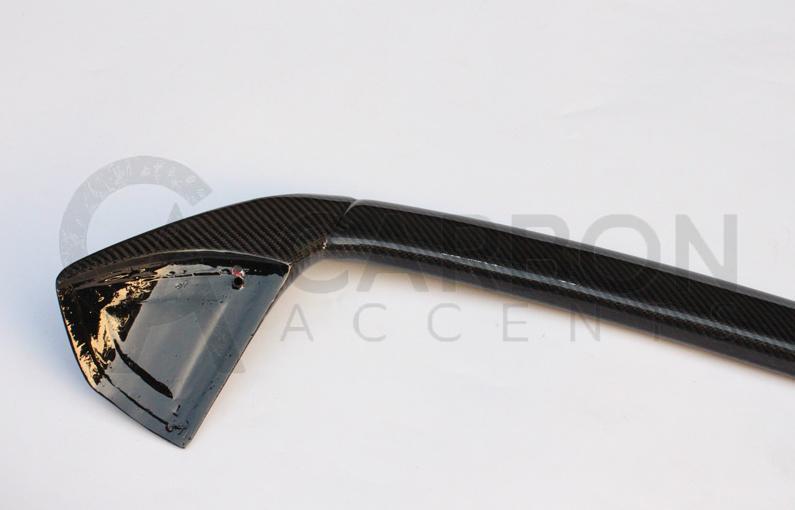 A black carbon fiber car spoiler rests on a white surface. Faint text in the background reads "CARBON ACCENT," partially obscured by the object's reflection.