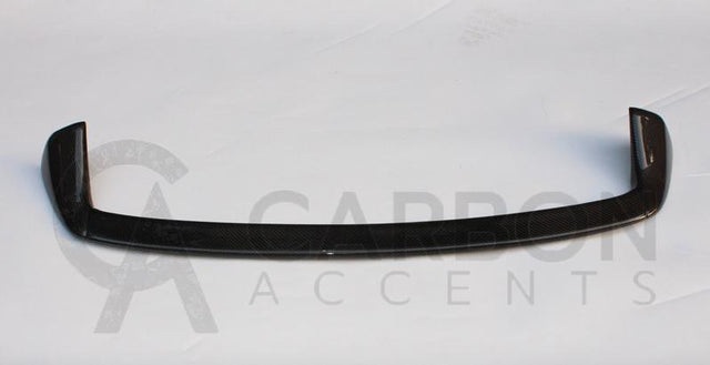 A carbon fiber car spoiler, curved and glossy, rests on a plain white surface. Text reads: "CARBON ACCENTS" with a partially visible stylized letter "A" behind the text.