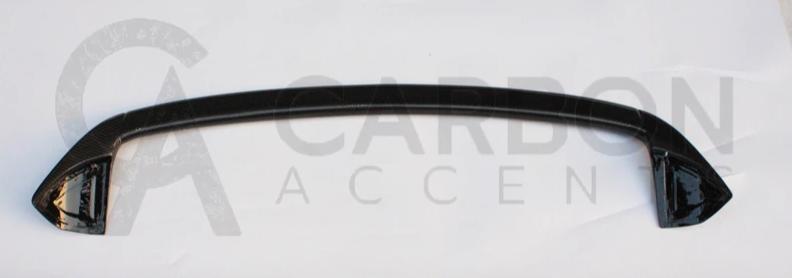 A carbon fiber car spoiler lies on a white surface. The text "CARBON ACCENTS" is faintly visible in the background.