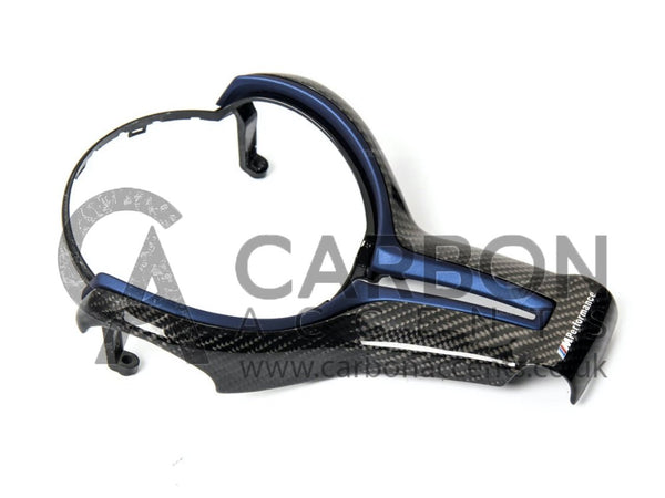 A carbon fiber car steering wheel trim with a blue accent rests on a plain surface. Text reads "M Performance" and a watermark "CARBON ACCENTS www.carbonaccents.co.uk" overlays the image.