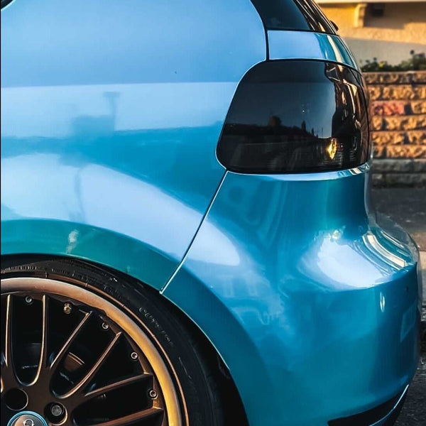 A blue car's rear end, showcasing tinted taillights and bronzed alloy wheels, is parked on a street. The background includes a building with warm-colored windows reflecting sunlight.