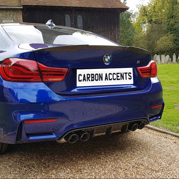 A blue BMW with the license plate "CARBON ACCENTS" is parked on a gravel path near a historic church building, surrounded by lush greenery and a small graveyard.