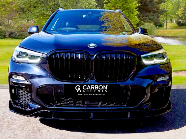 A black SUV is parked facing forward, showcasing its sleek design and prominent grille. Surrounding it are green trees and a pond. Text on the car reads, "CARBON ACCENTS www.CARBONACCENTS.co.uk".