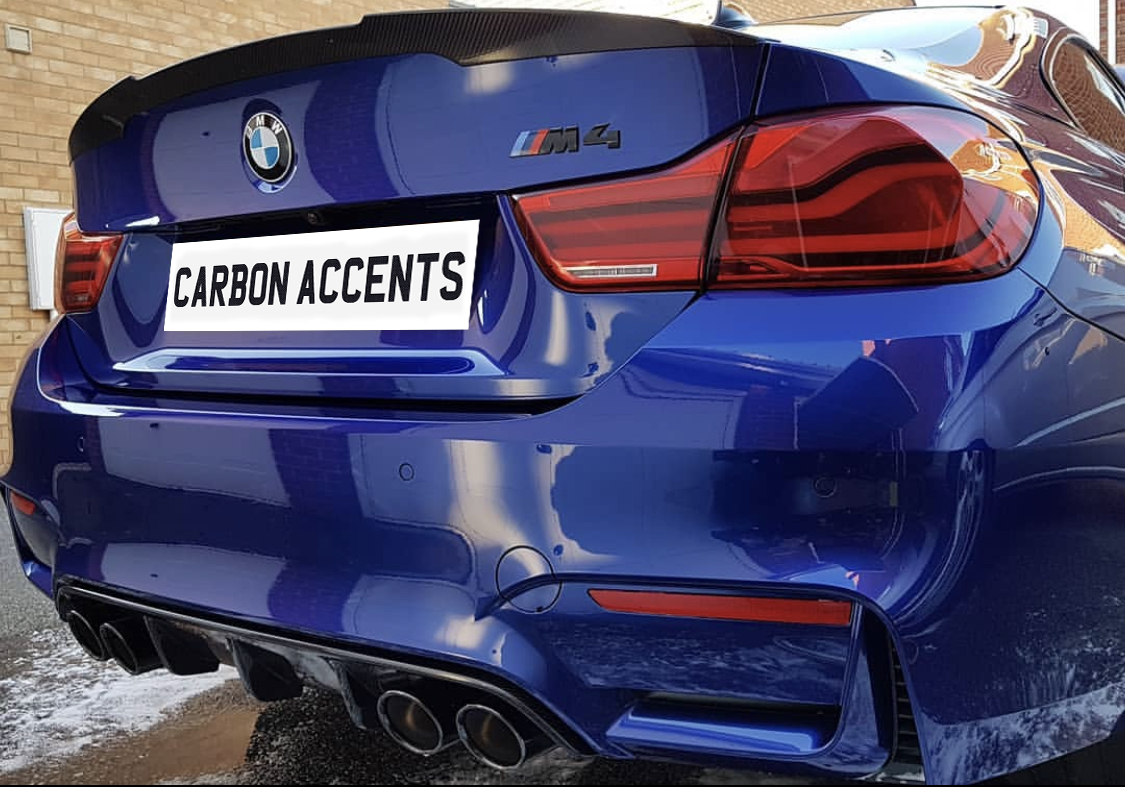 A blue BMW M4 rear view with quad exhausts and red taillights, parked beside a brick wall. The license plate reads "CARBON ACCENTS."