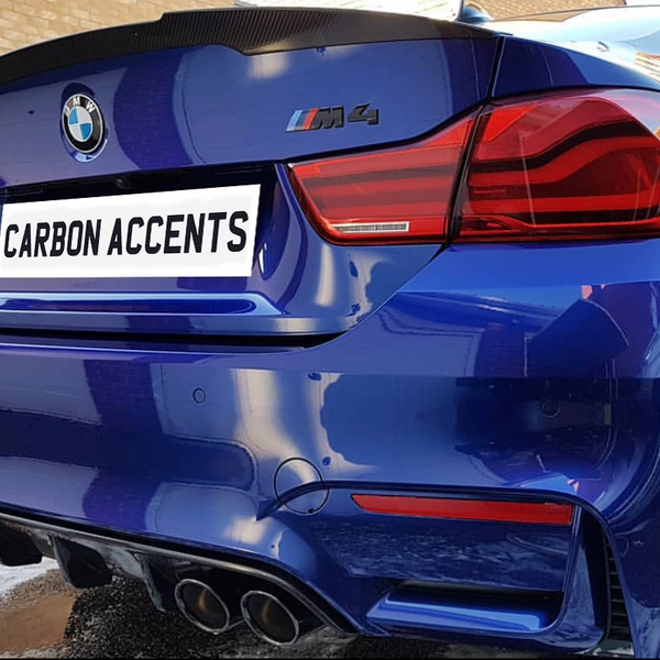 A blue BMW M4 rear view with quad exhausts and red taillights, parked beside a brick wall. The license plate reads "CARBON ACCENTS."
