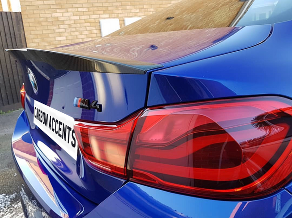 A blue BMW car with a carbon fiber spoiler is parked near a brick wall. The license plate reads "CARBON ACCENTS," and there's an "M4" badge visible.