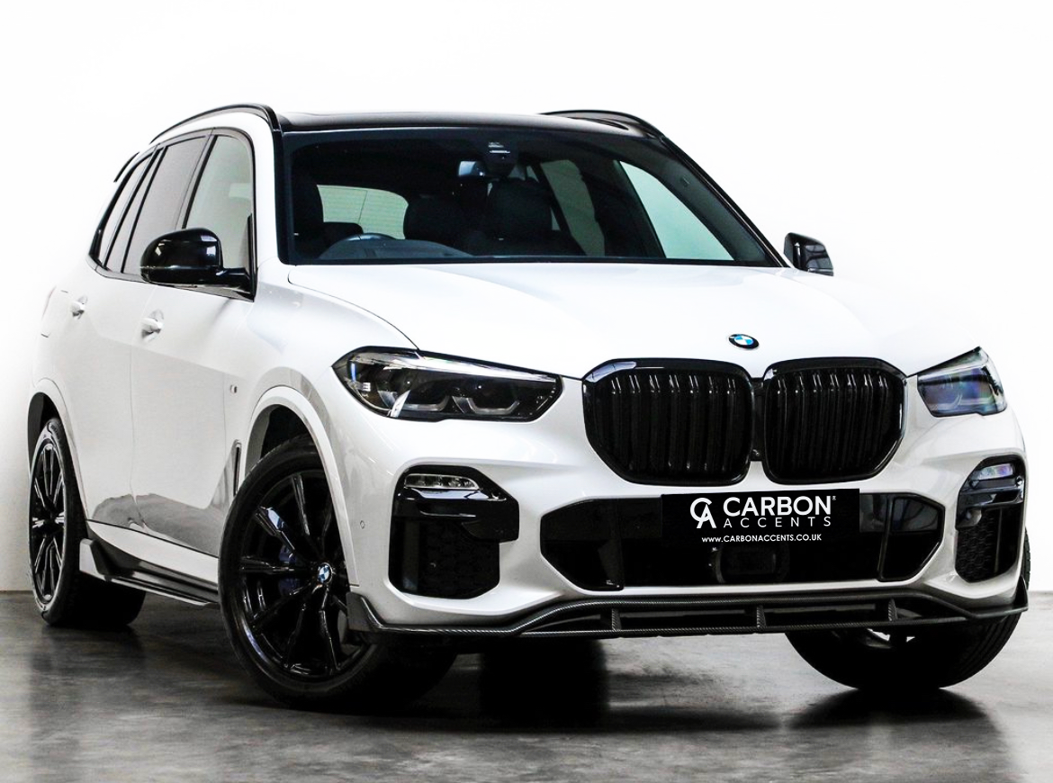 A white SUV with black accents is parked on a polished concrete floor in a studio setting. The front license plate reads, "CARBON ACCENTS www.CARBONACCENTS.CO.UK."