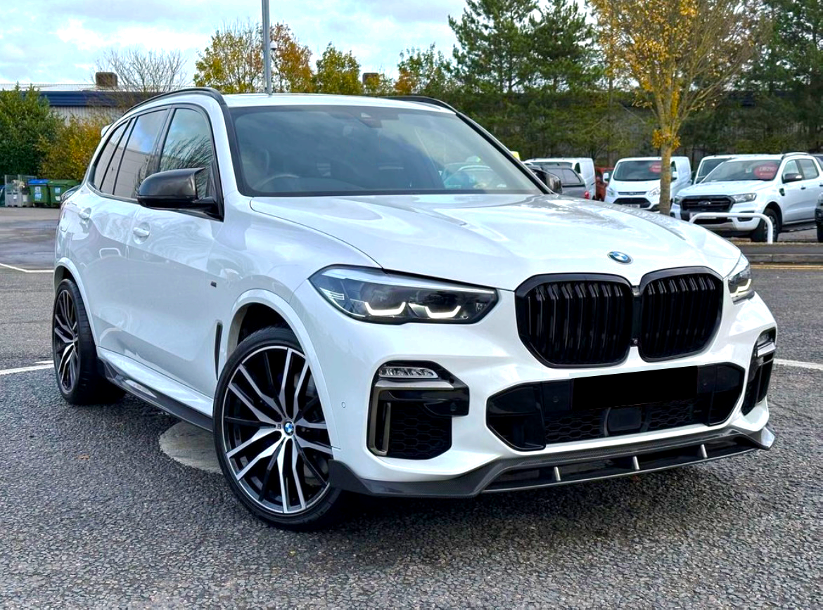 A white SUV is parked on a paved area, surrounded by trees and other vehicles in the background. It has a prominent black grille and large alloy wheels.