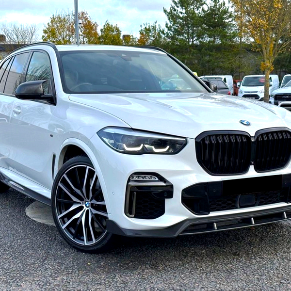 A white SUV is parked on a paved area, surrounded by trees and other vehicles in the background. It has a prominent black grille and large alloy wheels.