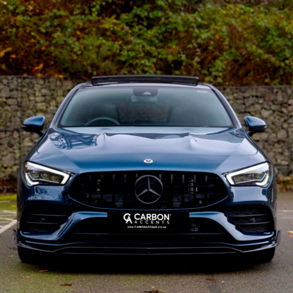 A blue car is parked in a paved area, featuring a prominent front grille with a logo. Behind it, a stone wall and lush greenery create a natural backdrop. Text: "CARBON ACCENTS" and "www.carbonaccents.co.uk".