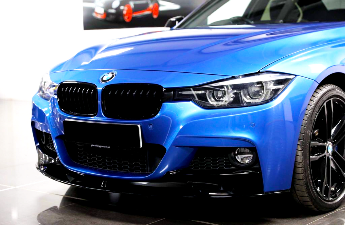 A blue BMW car is parked indoors, showcasing its sleek front grille, chrome logo, and shiny black wheels. The background features a blurred image of a car poster.