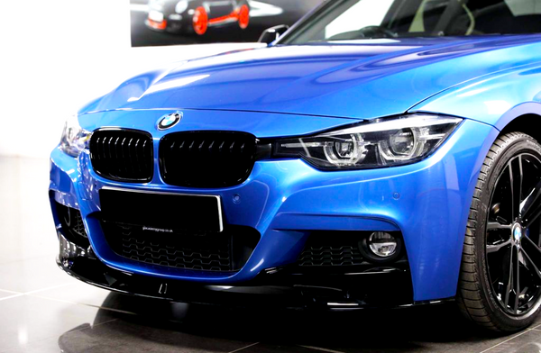 A blue BMW car is parked indoors, showcasing its sleek front grille, chrome logo, and shiny black wheels. The background features a blurred image of a car poster.