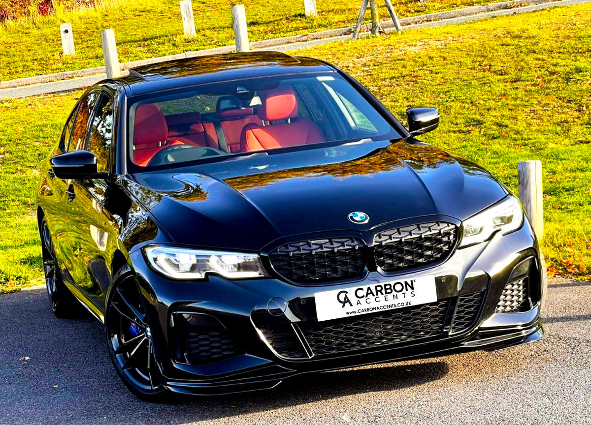 A black BMW sedan with red interior is parked on a paved road, surrounded by grassy areas. The license plate reads "CARBON ACCENTS, WWW.CARBONACCENTS.CO.UK."