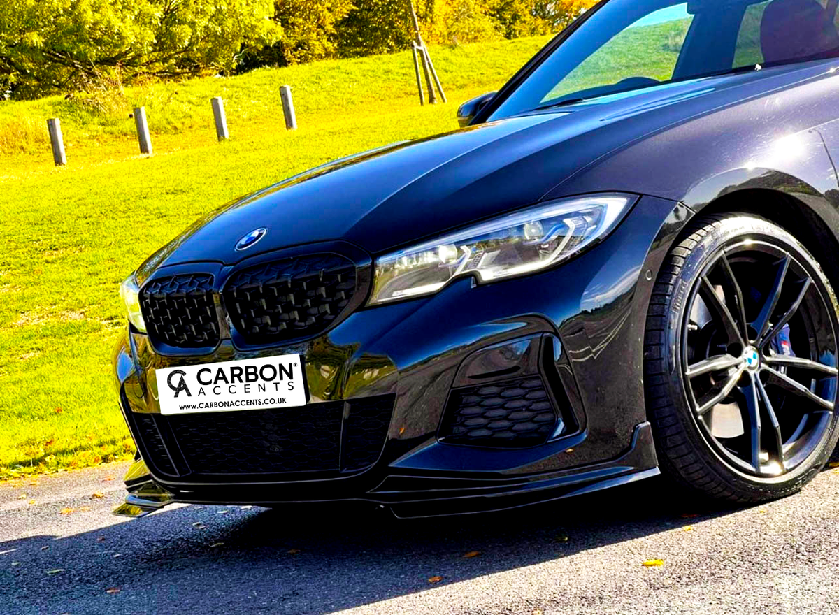 A black car with sleek lines is parked, featuring a "Carbon Accents" front license plate. It's on a paved path with a grassy field and trees in the background. Text: "CARBON ACCENTS www.carbonaccents.co.uk".