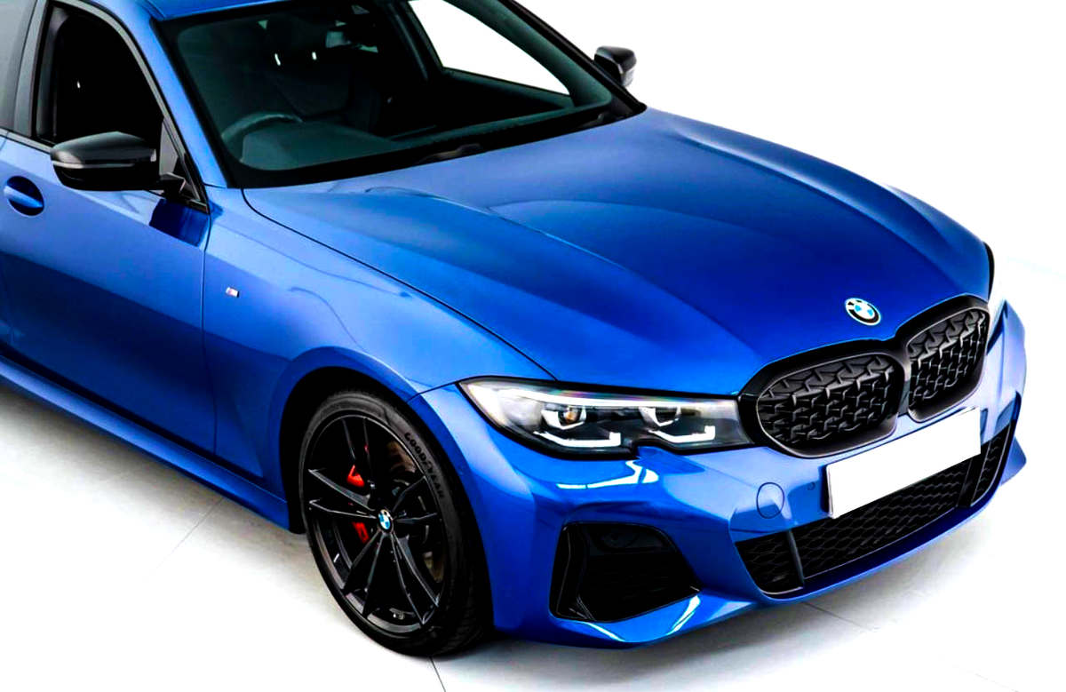 A metallic blue BMW car is stationary indoors, featuring a black grille, sporty headlights, and black wheels with red brake calipers. The environment is brightly lit, enhancing its sleek design.
