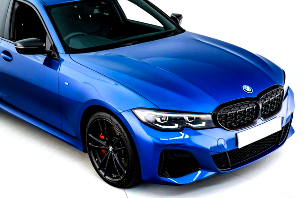 A metallic blue BMW car is stationary indoors, featuring a black grille, sporty headlights, and black wheels with red brake calipers. The environment is brightly lit, enhancing its sleek design.