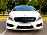White car with a black grille and circular emblem, parked in an outdoor setting surrounded by lush green trees and a clear sky. License plate text: 