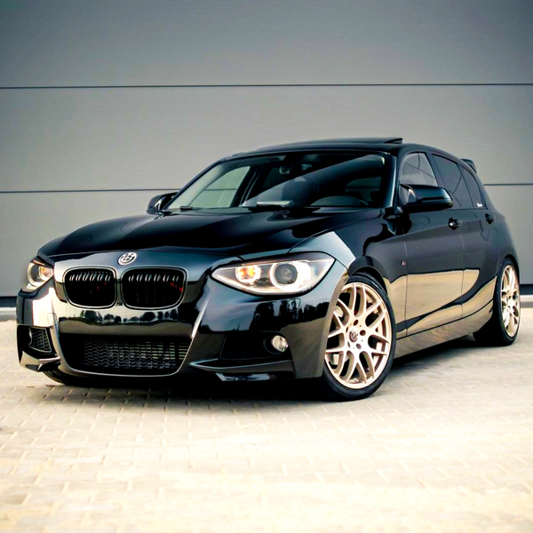A sleek black car with a sporty design is parked on a paved surface in front of a plain gray wall, featuring shiny alloy wheels and prominent front headlights.