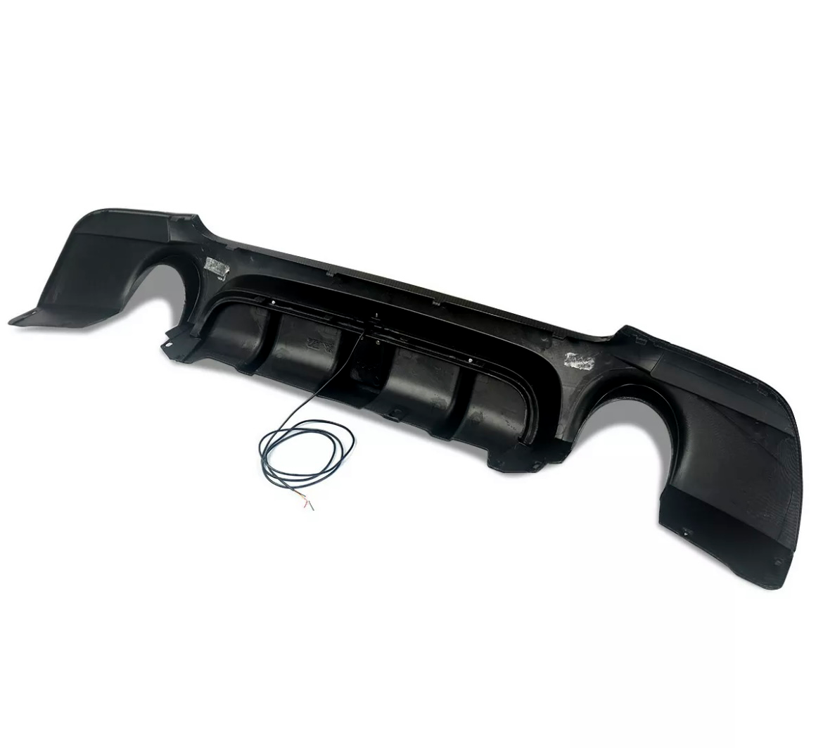 A black car rear diffuser, featuring aerodynamic fins, rests on a plain white surface with an attached cable coiled nearby.