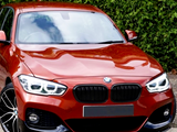 Red BMW car parked, facing forward, with headlights on; in front of a green hedge, on a paved surface.