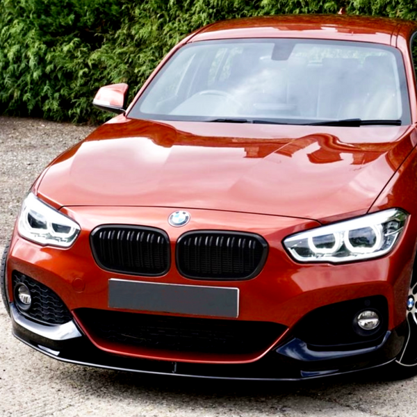 A red car with sleek headlights and a black grille is parked on a gravel surface, surrounded by green foliage.
