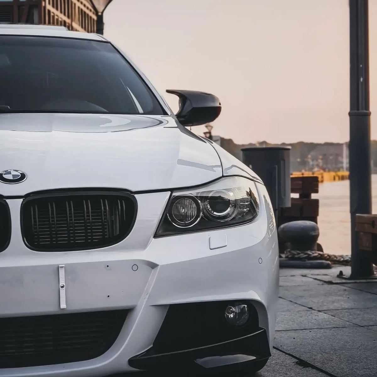 A white BMW car is parked, partially shown from the front. It features a sleek grille and headlights, located in a waterfront area with a calm atmosphere.