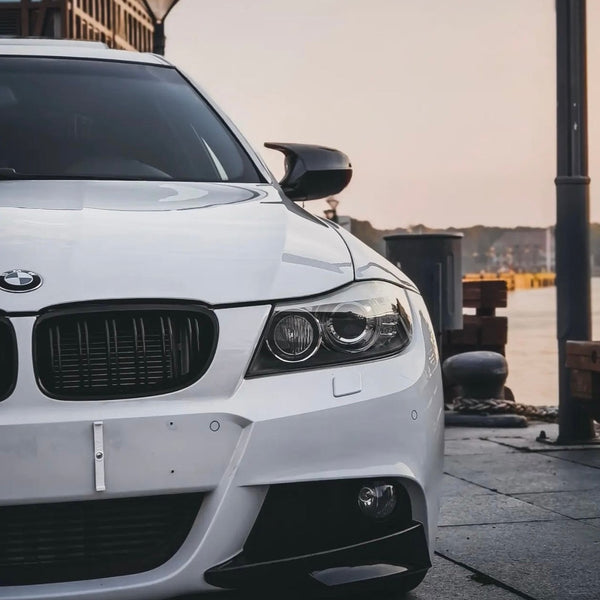 A white BMW car is parked, partially shown from the front. It features a sleek grille and headlights, located in a waterfront area with a calm atmosphere.