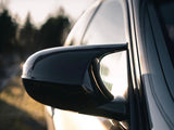 A sleek black side mirror reflects sunlight, attached to a modern car body with blurred trees and a road in the background, suggesting a serene outdoor setting.