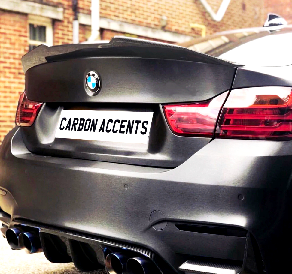 A rear view of a black BMW with notable carbon accents. The license plate reads "CARBON ACCENTS." The car is parked near a brick building.