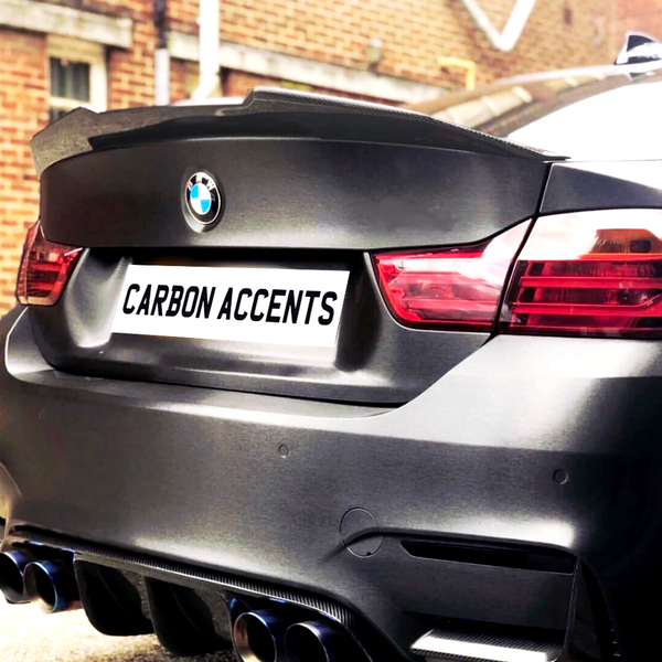 A rear view of a black BMW with notable carbon accents. The license plate reads "CARBON ACCENTS." The car is parked near a brick building.