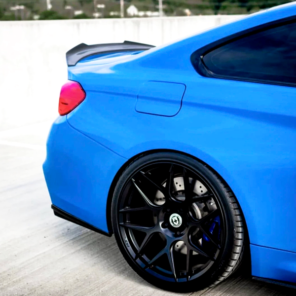 A blue sports car with sleek black wheels is parked on a concrete surface, featuring a prominent rear spoiler. The background shows a blurred landscape with greenery and buildings.