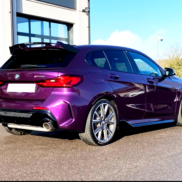 A purple BMW car is parked on a sunlit driveway beside a modern building with large windows. The surrounding landscape features green fields and a clear blue sky.