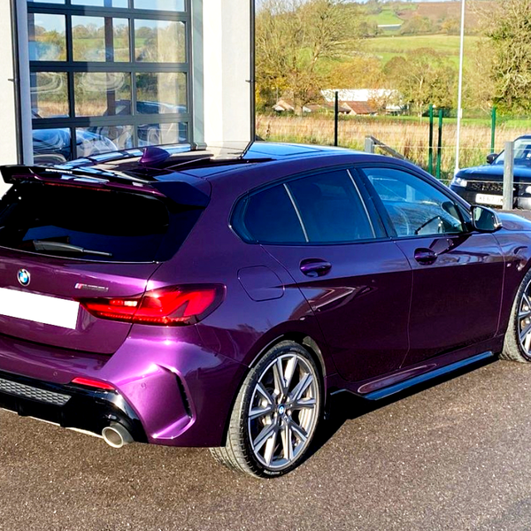 A purple hatchback car is parked on a paved surface beside a building with glass doors, with a scenic countryside landscape in the background.