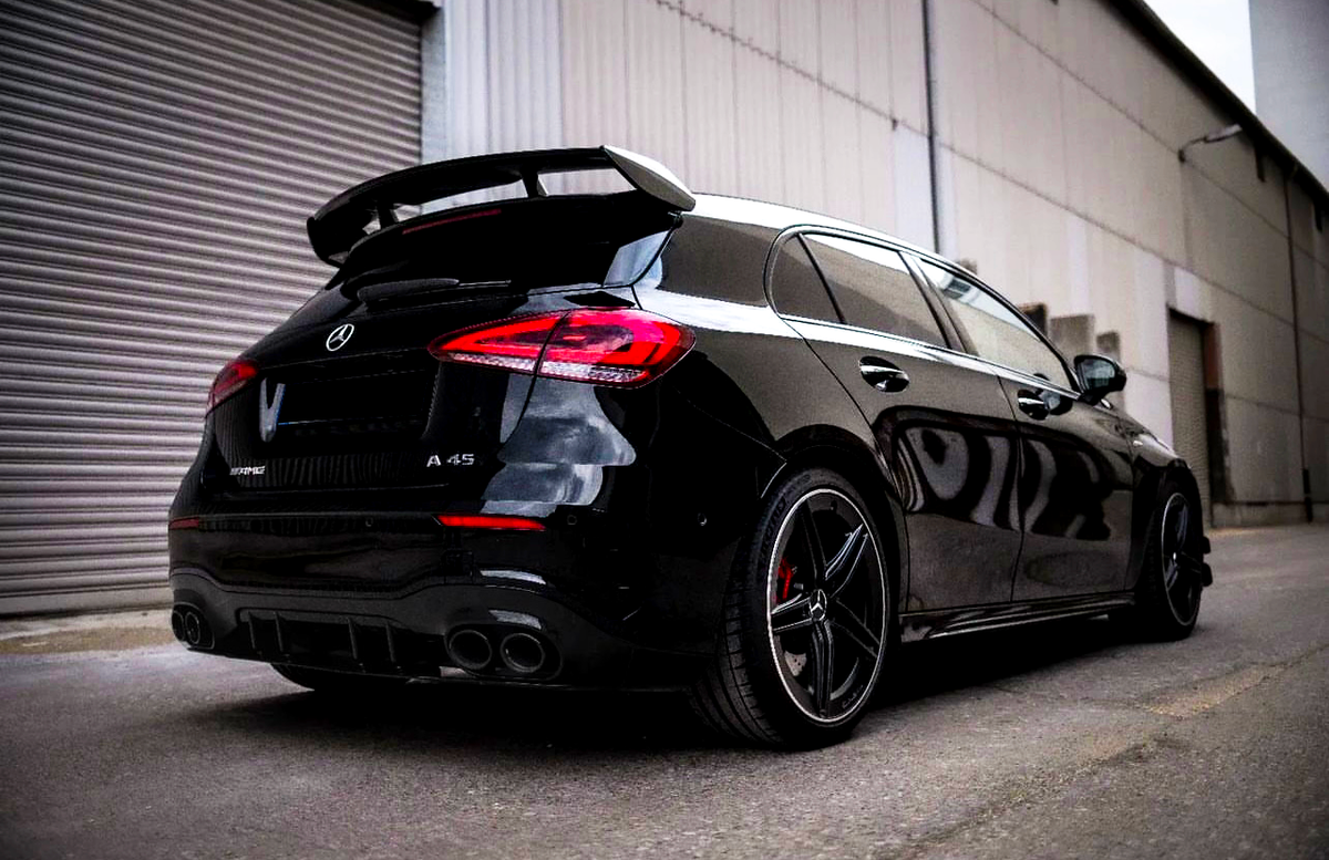 A black Mercedes-Benz A45 AMG parked, showing its rear design with dual exhausts and spoiler, in an industrial setting near a large metal shutter.