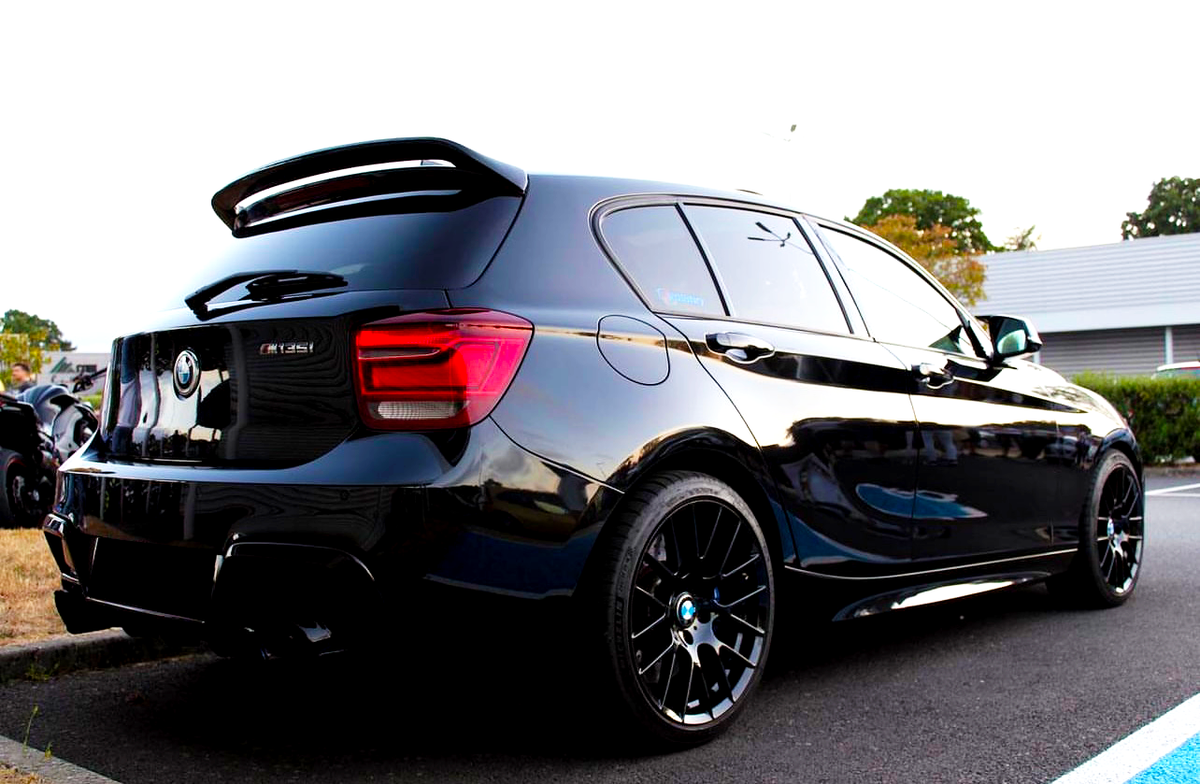 A black BMW 135i parked in a lot, featuring tinted windows, a rear spoiler, and glossy finish. Surrounding context includes greenery and a building.