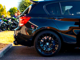 A black car with custom rims is parked beside a blue line in a lot. In the background, several motorcycles are parked near green bushes and a pole.