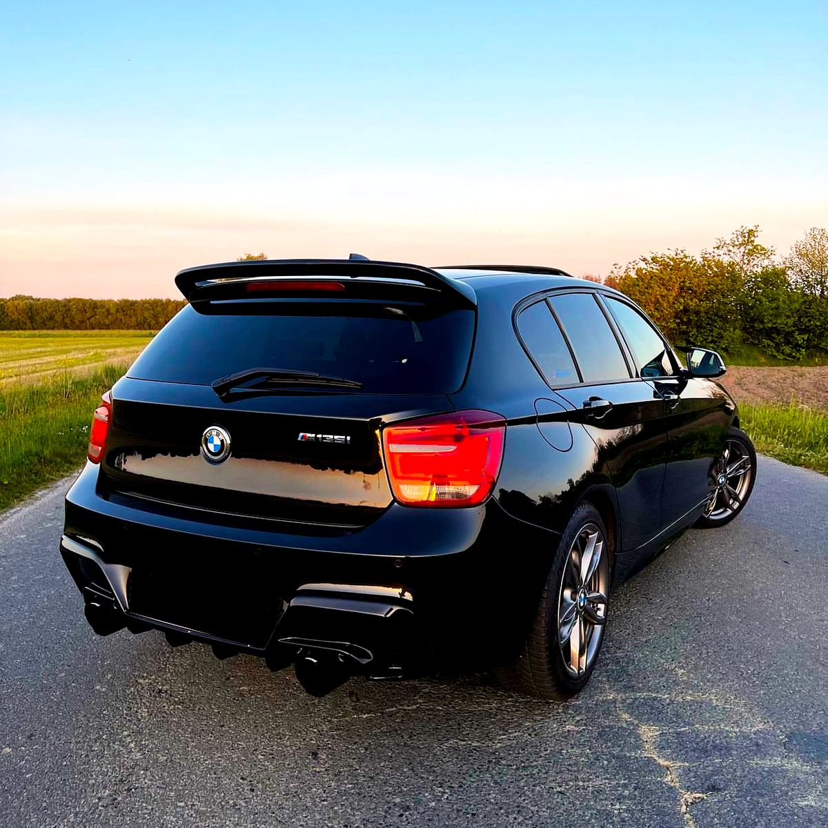 A black BMW M135i is parked on a deserted rural road during sunset, surrounded by green grass and trees under a clear blue sky.