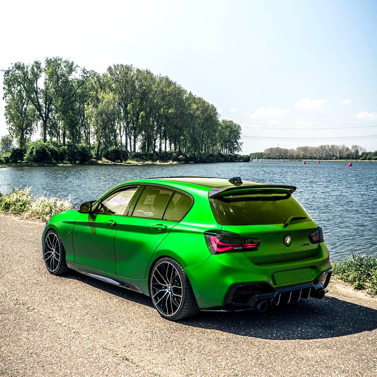 A vibrant green car is parked on a paved road beside a calm river, with a row of tall trees lining the opposite riverbank under a clear blue sky.