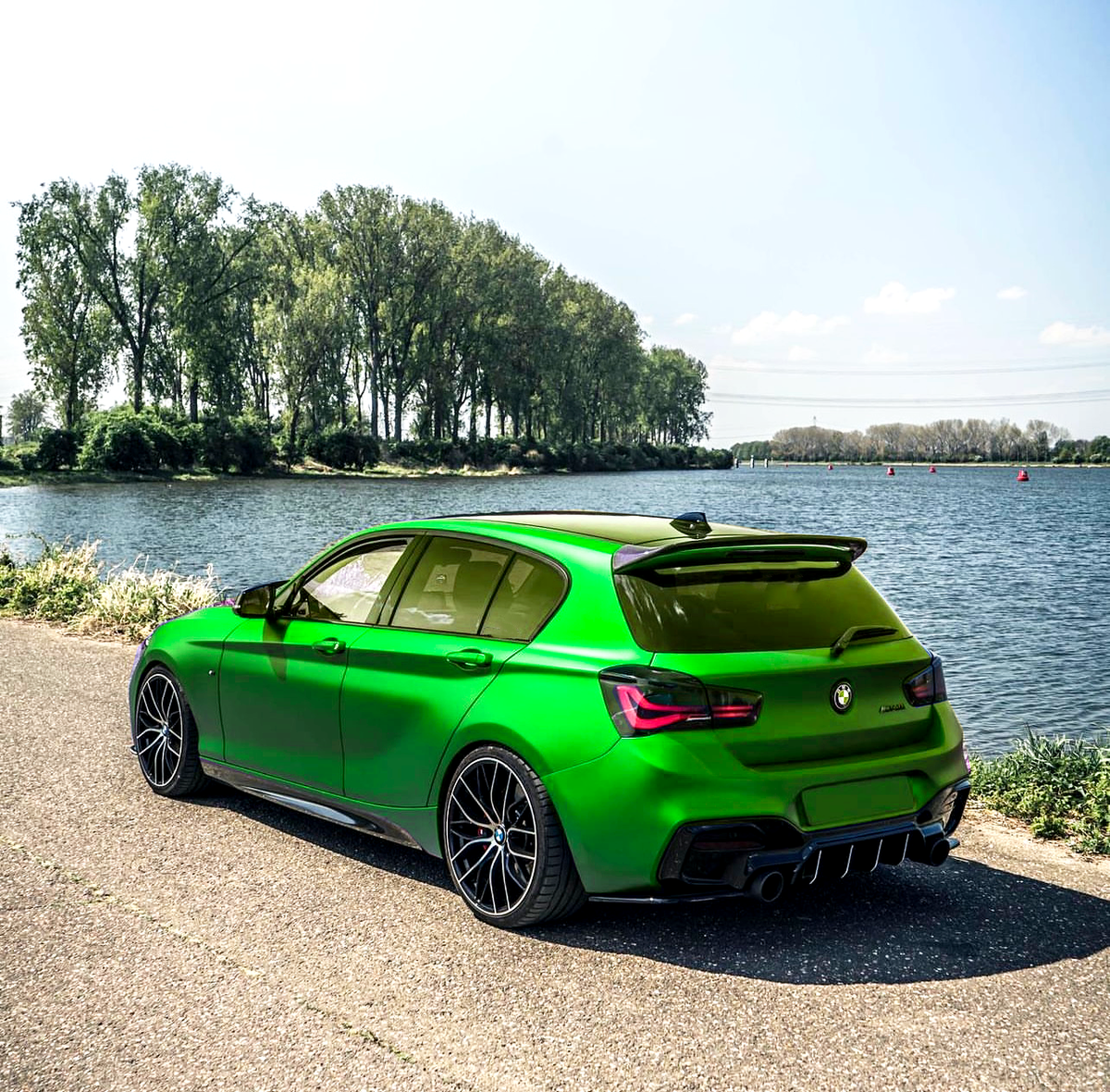 A vibrant green car is parked on a paved road beside a calm river, with a row of tall trees lining the opposite riverbank under a clear blue sky.