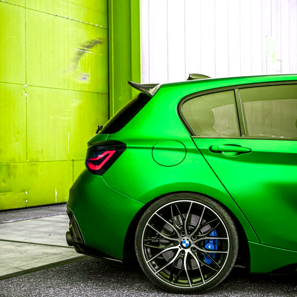 Green sports car parked near a yellow and white wall. The image focuses on the rear wheel, which features a blue brake caliper and distinctive alloy design.