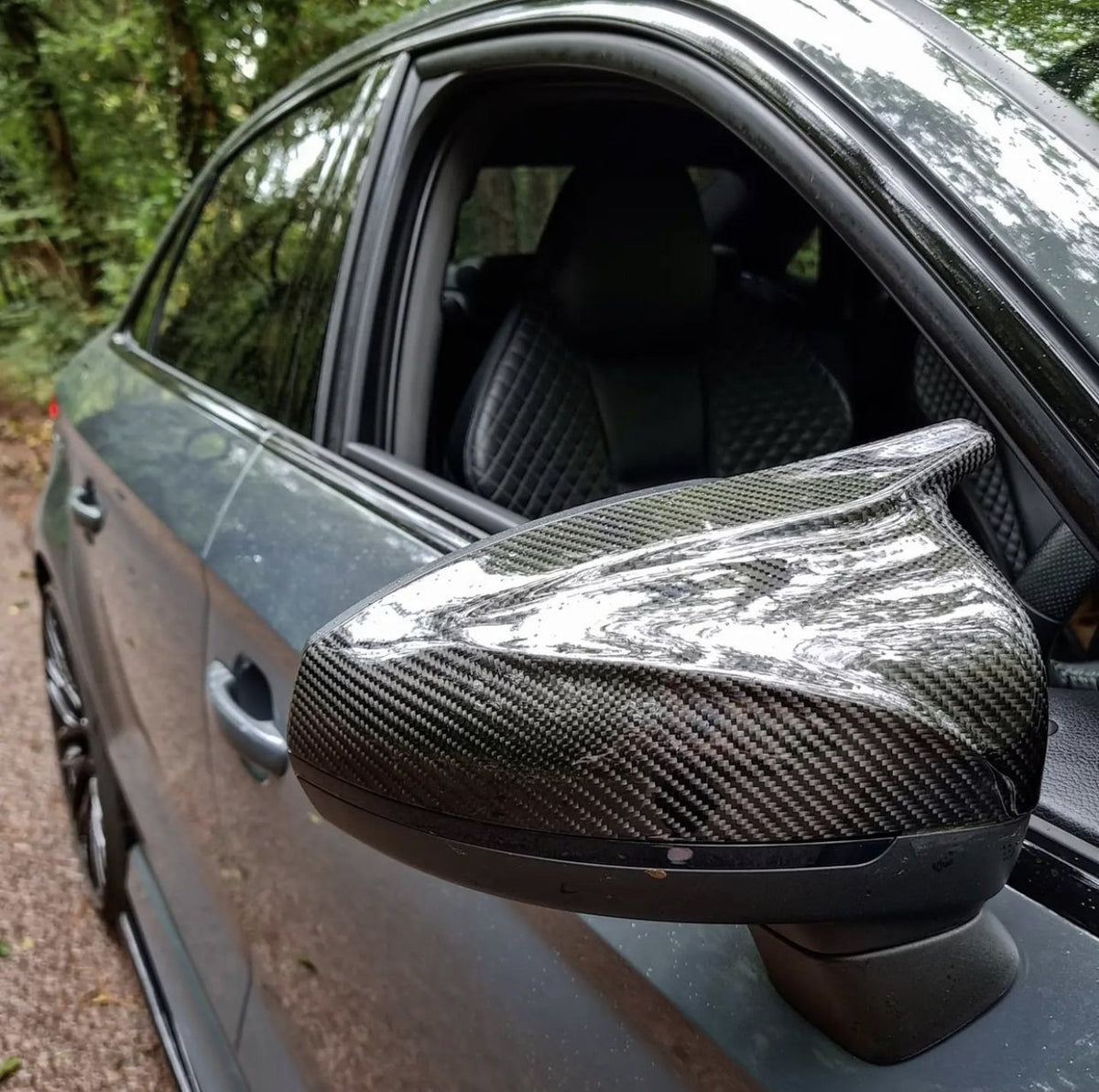 A carbon fiber side mirror on a sleek car reflects the forest canopy above, with partially visible quilted leather seats inside the vehicle. The car is parked on a forest path.