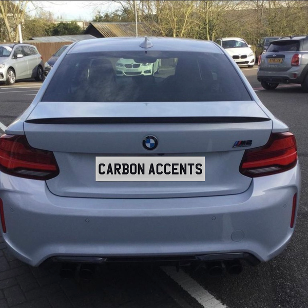 A silver BMW car is parked, displaying the text "CARBON ACCENTS" on its rear plate. It's situated in a parking lot with various other vehicles nearby.