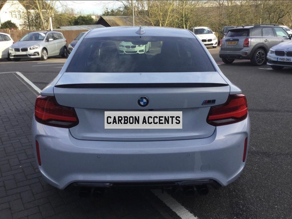 A silver BMW car is parked, displaying the text "CARBON ACCENTS" on its rear plate. It's situated in a parking lot with various other vehicles nearby.
