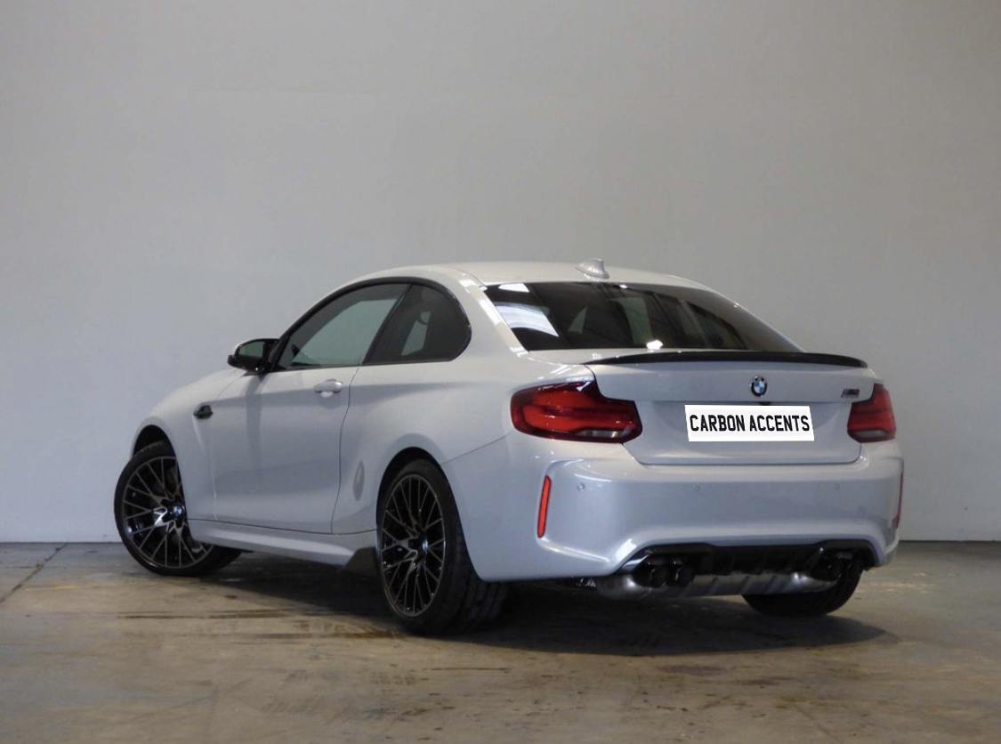 A silver BMW coupe parked indoors on a concrete floor, featuring black wheels and a visible “CARBON ACCENTS” sticker on its rear.