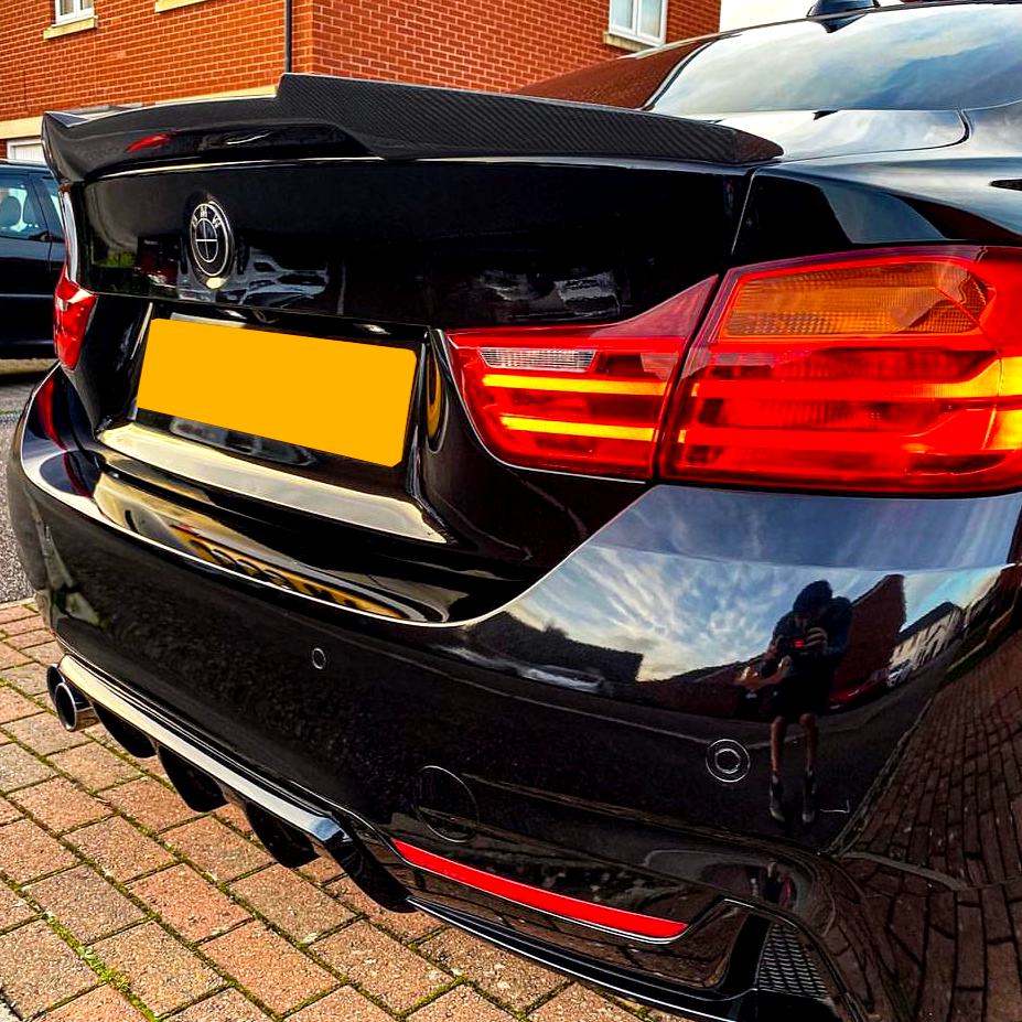 A black car with a spoiler is parked on a brick driveway. The rear shows distinct red taillights and a logo resembling Toyota’s. Reflections on the surface include sky and houses.