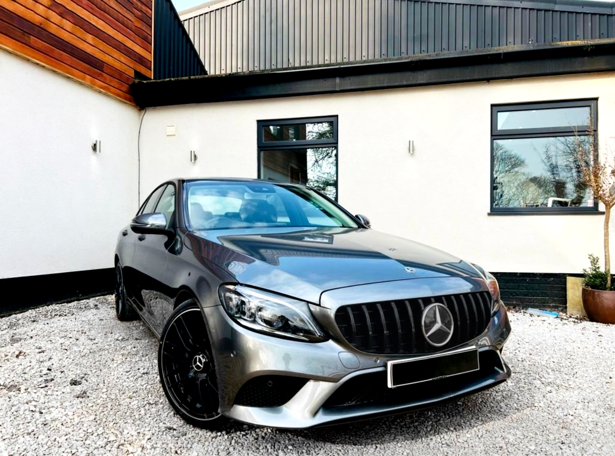 A silver Mercedes-Benz car is parked on gravel in front of a modern house with white and wooden exterior walls. The car's sleek design and black rims are prominent.