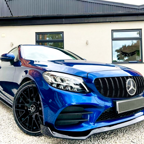 Blue Mercedes car parked on gravel, showcasing black wheels and grille. The background features a modern house with white walls, wooden paneling, and large windows.